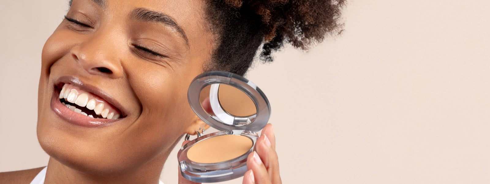 Model smiling with eyes closed holding 4-in-1 Pressed Powder against cheek - against a beige tan background