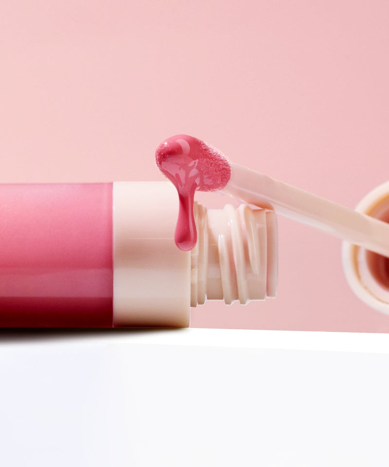 Glossy Lip Tint close up picture against light pink background and light colored surface with lip brush applicator dripping lip tint oil