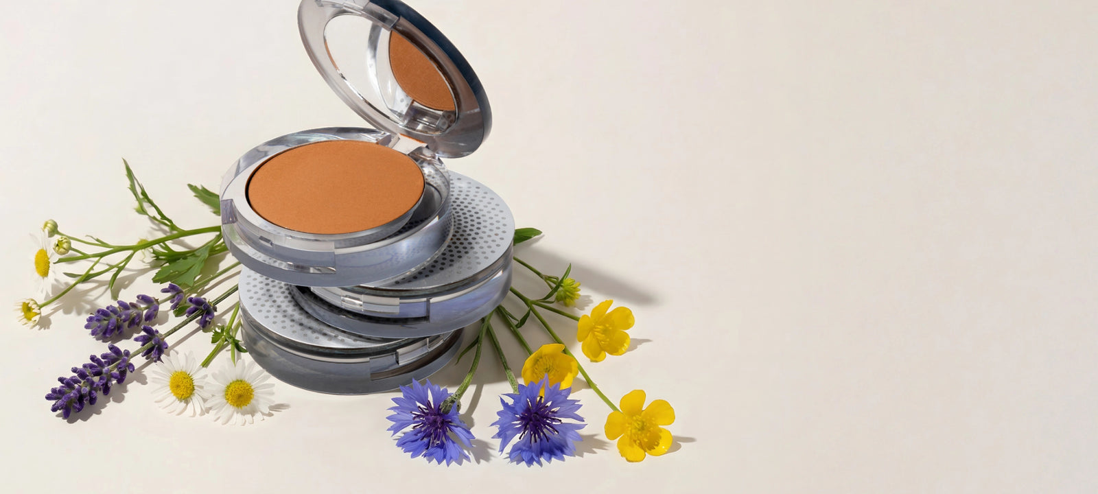 For Mom, with Love - Save up to $25: Stack of 3 4-in-1 Pressed Mineral Makeup products surrounded by yellow and purple flowers against tan background (desktop)