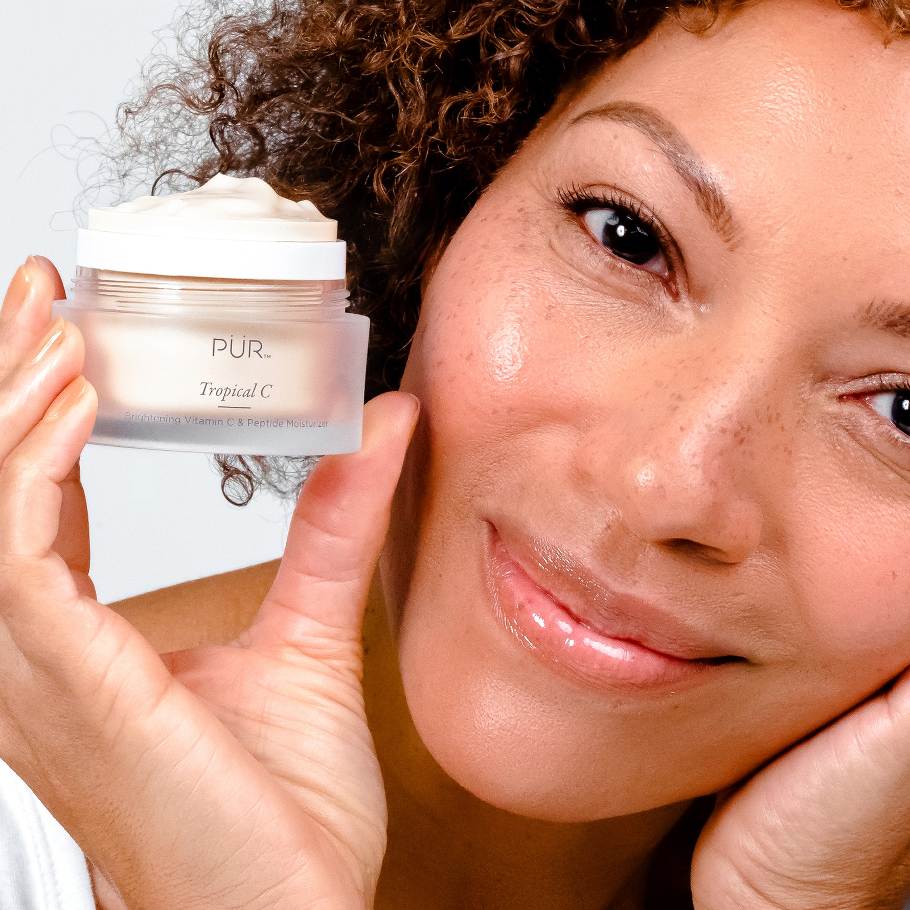 Model holding PÜR Tropical C Brightening Vitamin C & Peptide Moisturizer, showing glowing, hydrated skin and radiant complexion