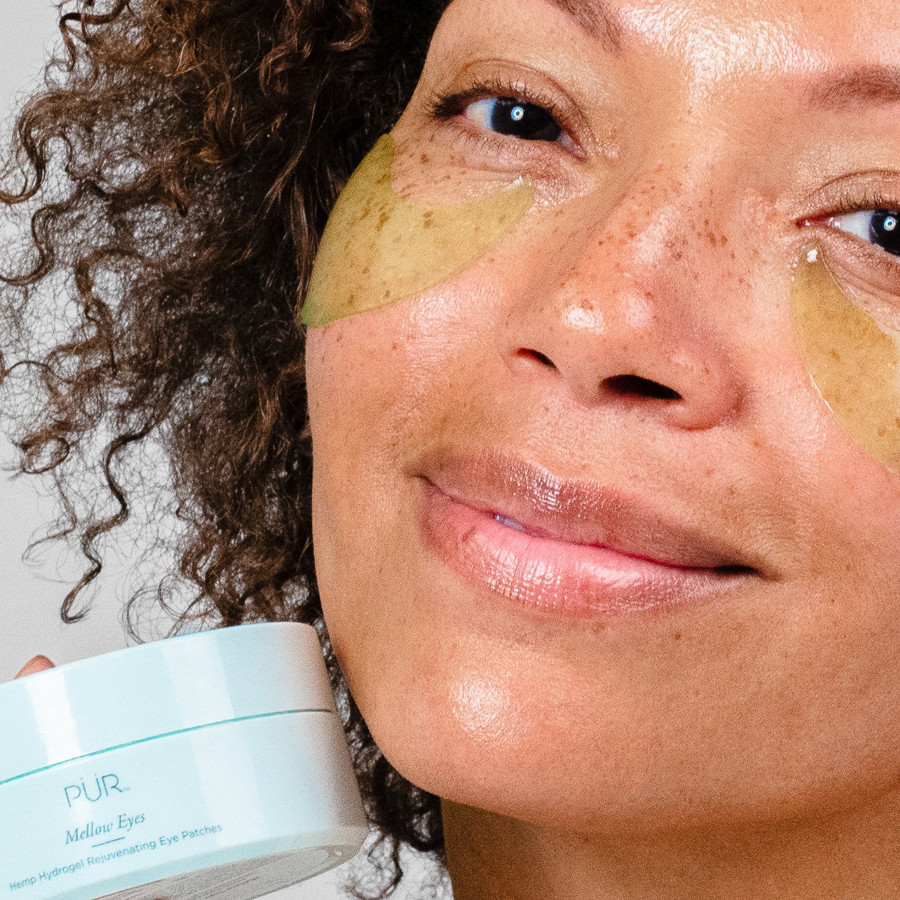 Woman wearing Mellow Eyes hydrogel eye patches while holding the product jar, showing refreshed under-eye glow.